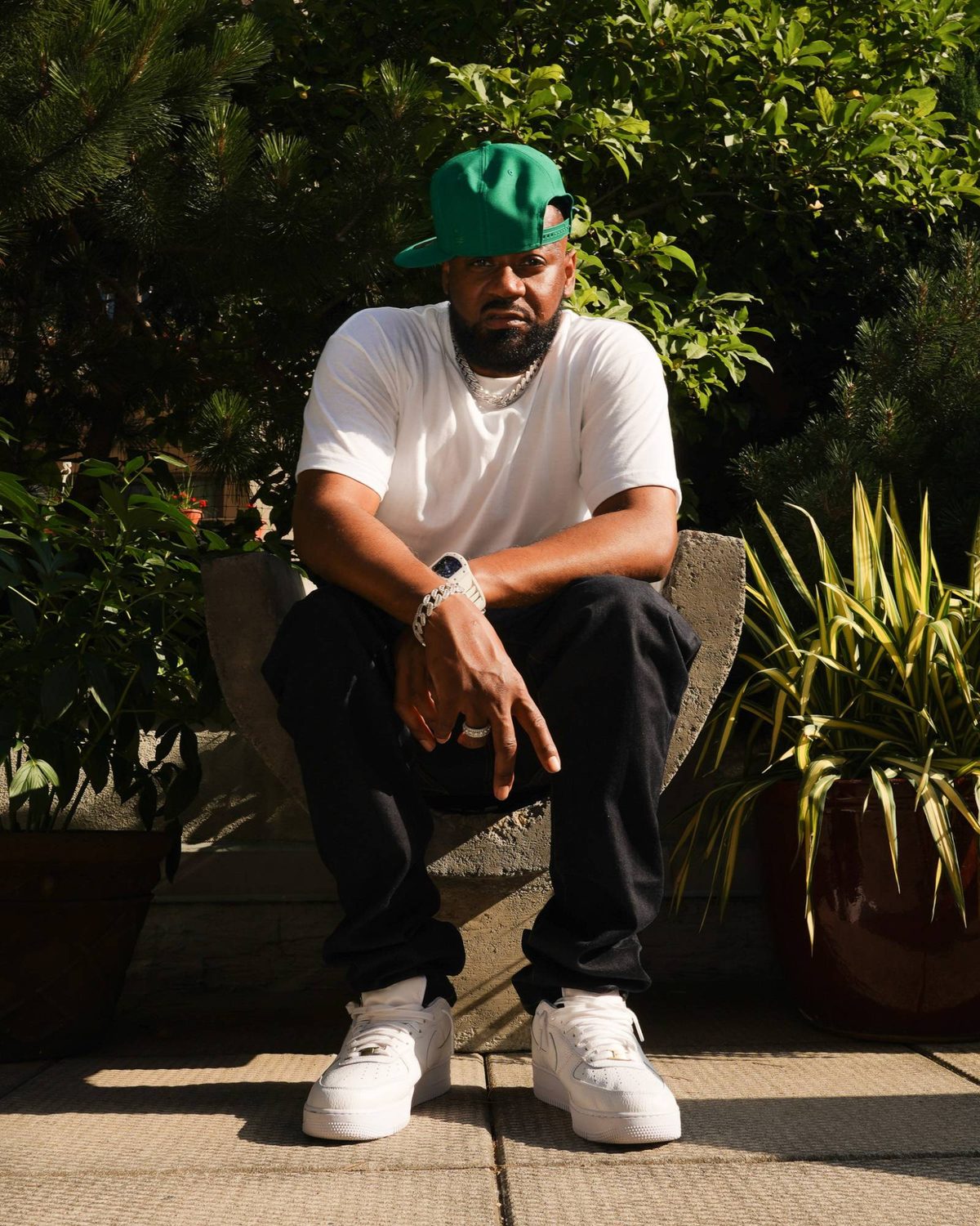 Ghostface Killah seated on a stone chair in a rooftop garden, green cap and white tee, in late sun