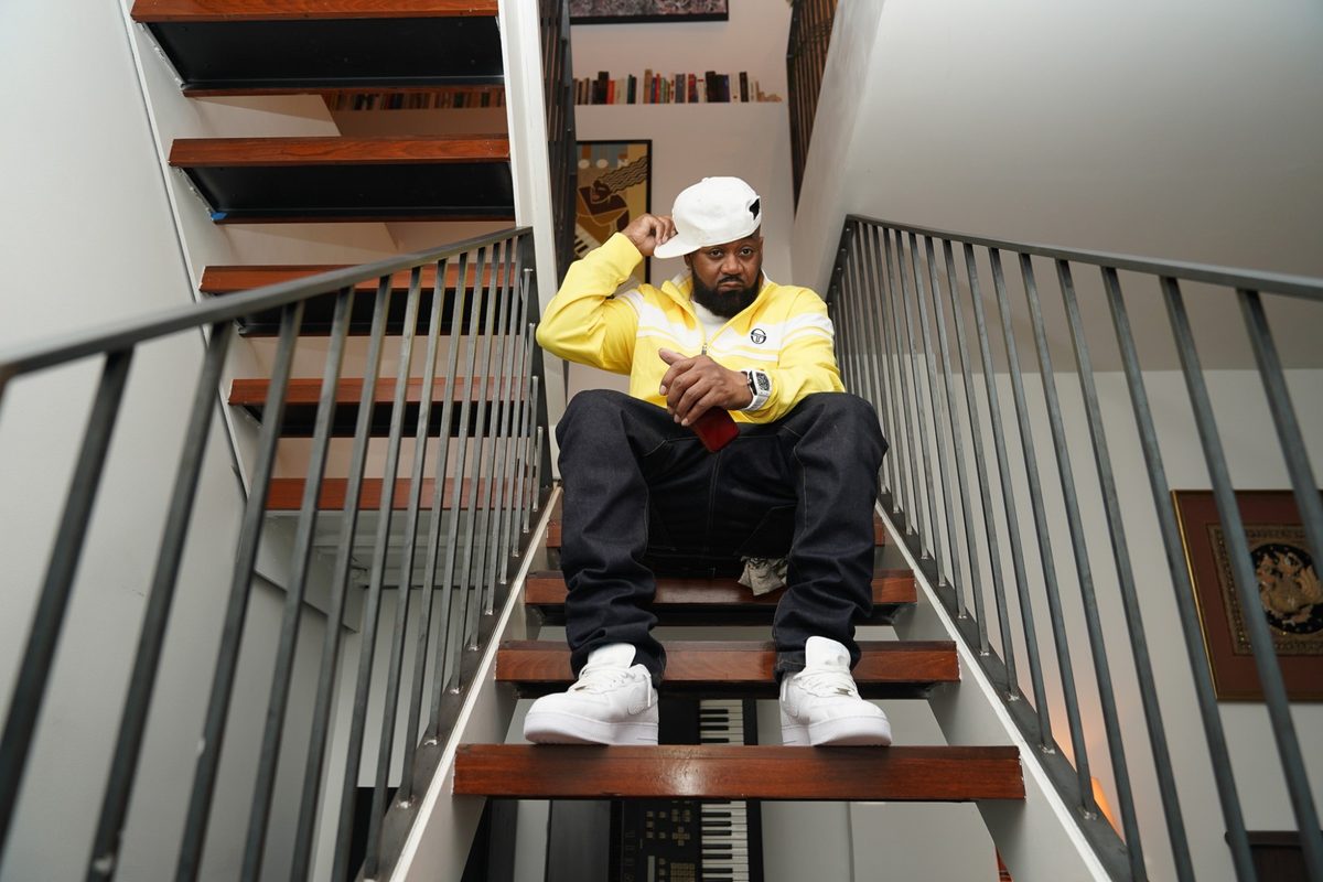 Ghostface Killah on an open staircase in a yellow track jacket, touching the brim of a white cap