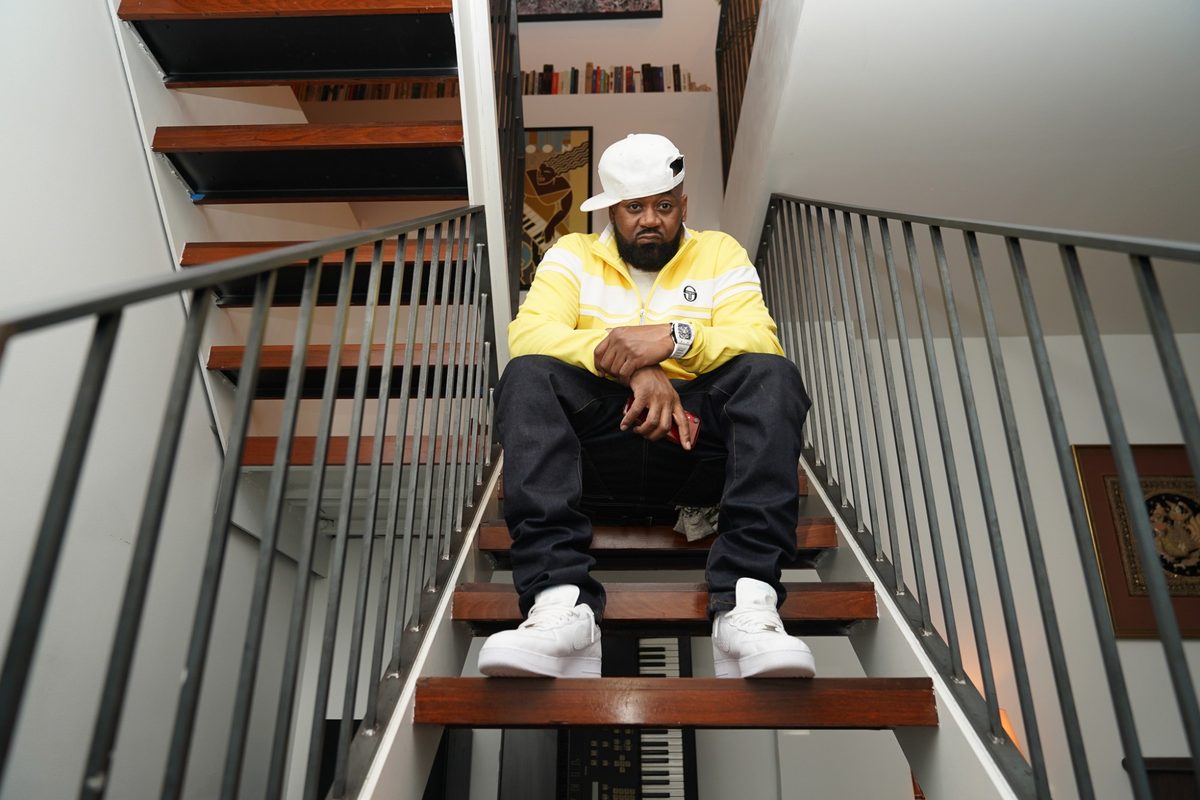Ghostface Killah seated on the stairs in a yellow track jacket, arms crossed, looking down the lens
