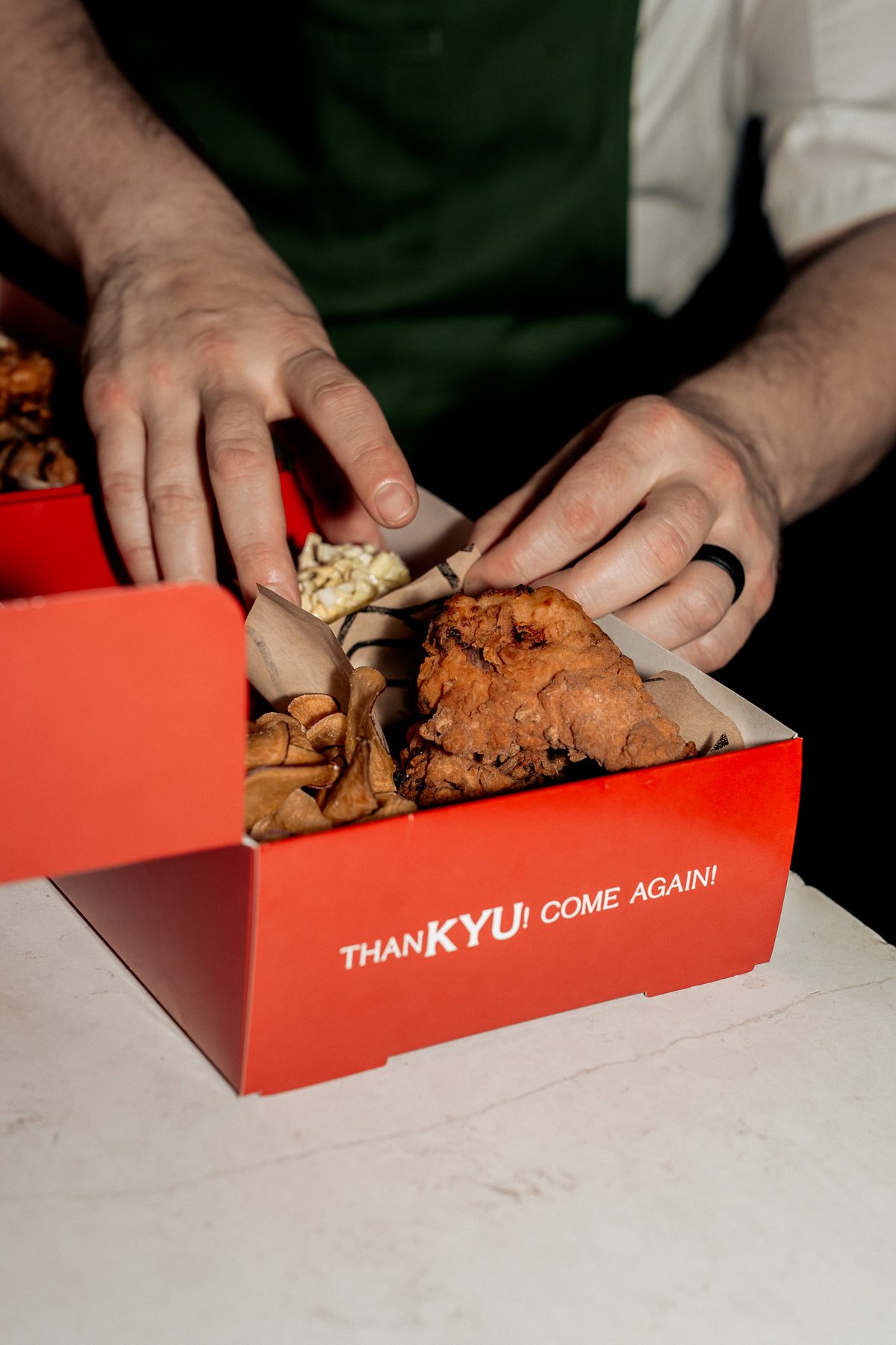 A cook's hands setting fried chicken into a red KYU takeout box printed “ThanKYU! Come Again!”