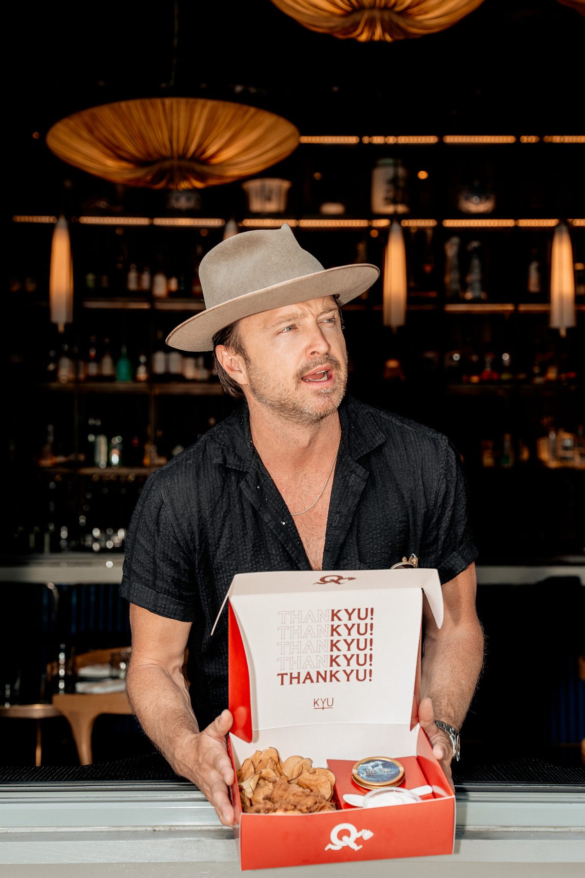 Aaron Paul smiling and presenting an open ThanKYU box at the bar