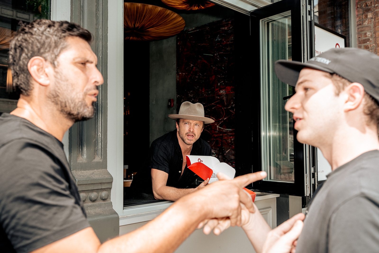 Two men talking outside the takeout window while Aaron Paul works behind it
