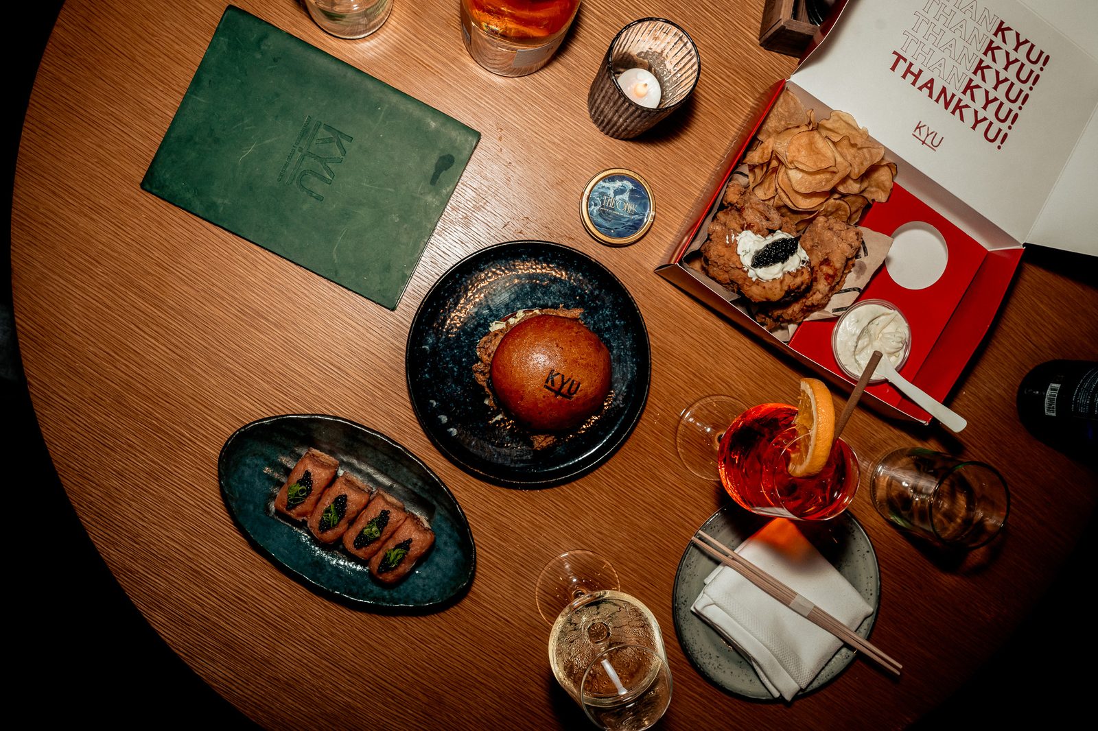 Overhead of the table: the ThanKYU box, a chicken sandwich, cocktails and small plates